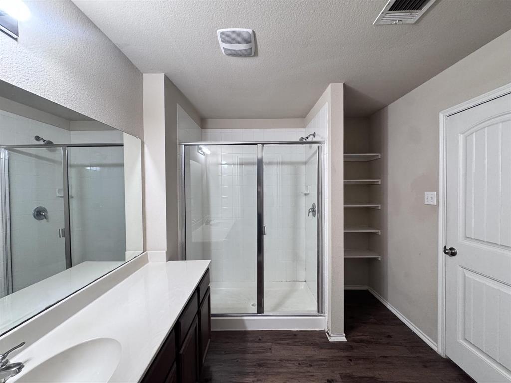 Full-width wall mirror, wood-finish flooring, glass-enclosed shower with tile surround, solid-surface vanity countertop, and built-in wall shelving
