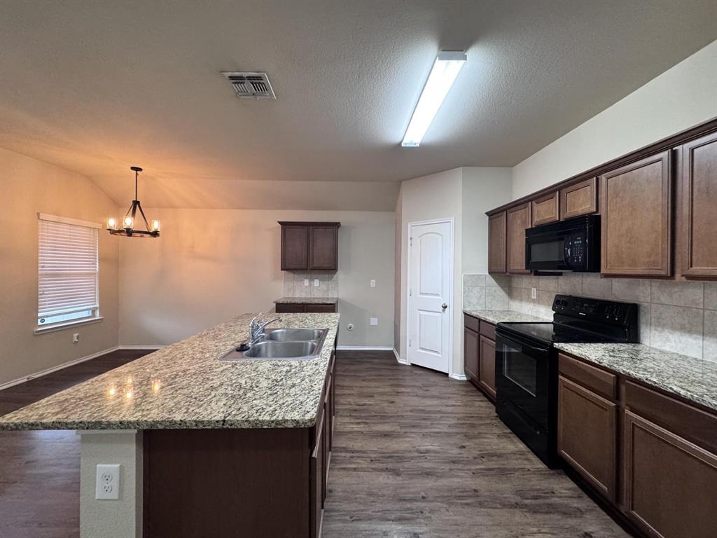 Kitchen featuring an expansive stone countertop island with a dual-basin stainless steel sink, wood cabinetry, stone-tiled backsplash, and wood-finish flooring
