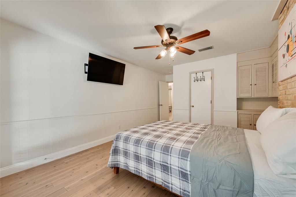 Wood-finish flooring, ceiling fan with integrated lighting, wall-mounted cabinetry with louvered doors, and brick accent wall