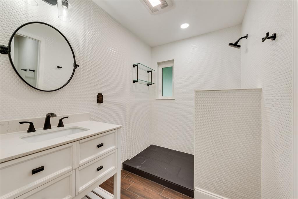 White vanity featuring a stone-surface countertop, circular mirror with matte black frame, matte black plumbing fixtures, penny-tile wall cladding, and walk-in shower with dark tile flooring
