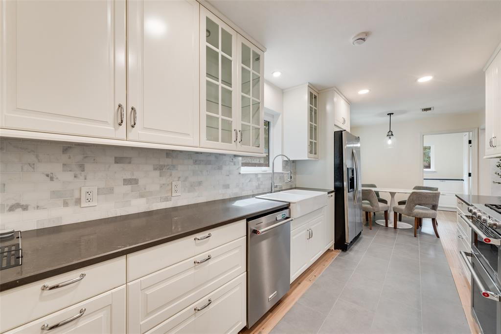 Kitchen featuring white cabinetry with glass-front uppers, solid-surface countertops, stone-tile backsplash, stainless steel appliances, and a farmhouse-style apron sink