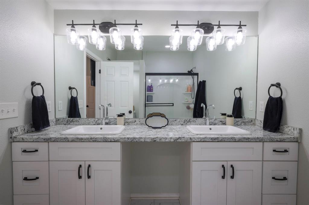 Dual-sink vanity featuring stone-finish countertops, white shaker-style cabinetry, matte black hardware, and double multi-bulb vanity lighting