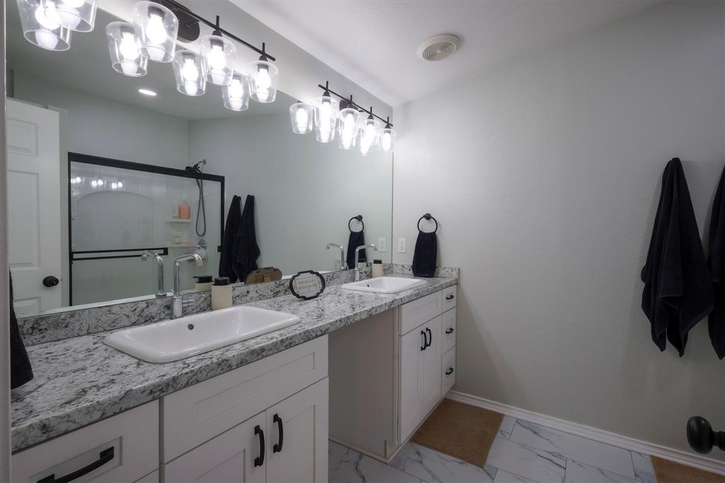 Dual-sink vanity with stone-finish countertops, undermount rectangular basins, matte black hardware, and wall-to-wall mirror with multi-bulb industrial-style lighting