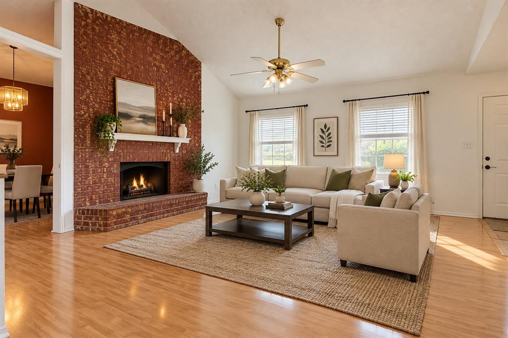 Living room featuring vaulted ceilings, wood-finish flooring, floor-to-ceiling brick fireplace, raised hearth, and brass-finish ceiling fan with integrated lighting Living room featuring vaulted ceilings, wood-finish flooring, floor-to-ceiling brick fireplace, raised hearth, and brass-finish ceiling fan with integrated lighting