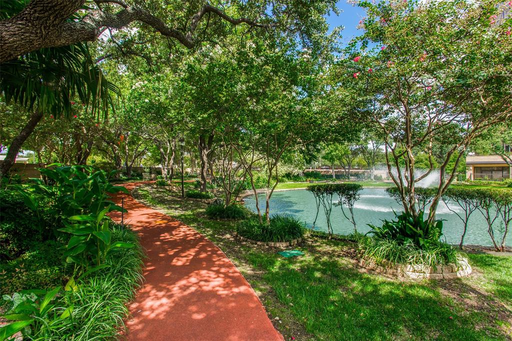 Landscaped grounds featuring a winding paved path, mature shade trees, stone-edged garden beds, and a decorative pond with a water feature