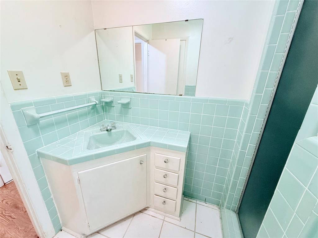 Corner vanity featuring a recessed basin, integrated tile countertop, and white cabinetry, accented by light green square wall tiling, a built-in towel rack, and a large wall-mounted mirror