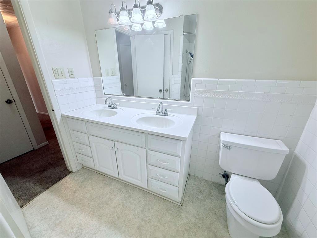 Double-sink vanity featuring a solid surface countertop, white shaker-style cabinetry, a wall-mounted mirror, and tile wainscoting
