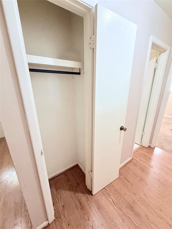 Storage closet featuring white-finish interior shelving, a metal rod, and a white swing door with a metallic knob, complemented by wood-finish flooring