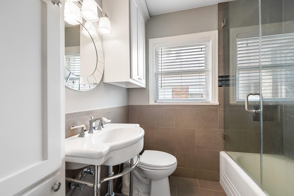 Pedestal sink with polished metal hardware, circular vanity mirror, built-in white wall cabinetry, neutral tile wainscoting, and a glass-enclosed shower-tub combination