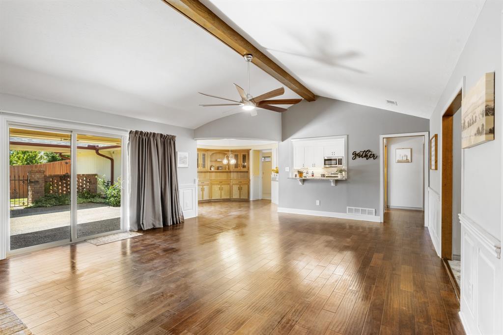 Spacious and inviting living area featuring rich wood flooring, vaulted ceilings with a striking exposed wood beam, and abundant natural light. The open layout flows seamlessly into the kitchen and dining areas, while French doors provide easy access to the covered outdoor living space.