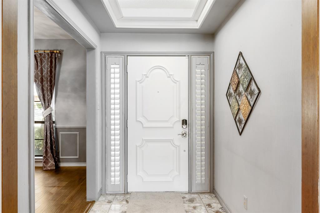 Welcoming entry foyer featuring a beautifully detailed front door flanked by plantation shutters and enhanced by a skylight for natural light. Crisp white trim and neutral finishes create a bright, polished first impression with a seamless view into the formal dining area.