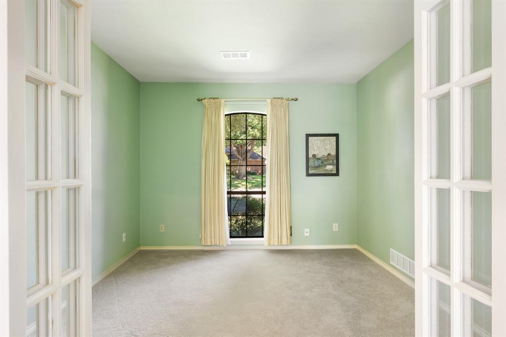 Light-filled private office featuring French doors, soft green walls, plush carpeting, and a charming arched window overlooking the front yard. The inviting space offers a peaceful setting for a home office, study, or flex space.