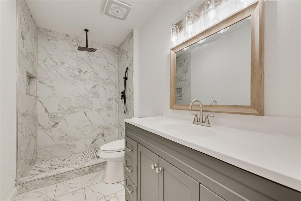 Bathroom featuring a walk-in shower with stone-pattern tile surround, recessed wall niche, matte black rainfall showerhead, gray shaker-style vanity with white solid-surface countertop, and wood-framed mirror
