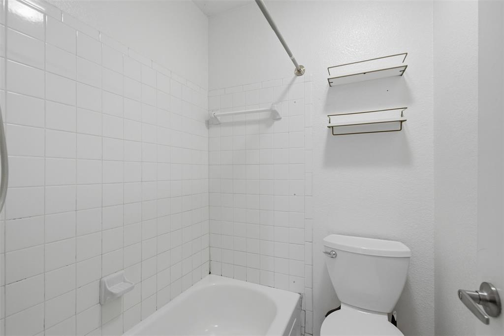 Full-height white square tile tub surround, built-in soap dish, shower curtain rod, wall-mounted storage shelving, and standard porcelain toilet