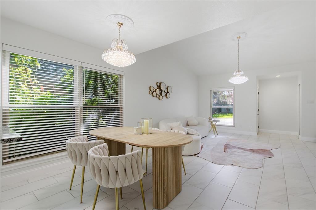 Open-concept living space featuring vaulted ceilings, light-toned tile flooring, dual crystal chandeliers with matching ceiling medallions, and large windows with horizontal blinds
