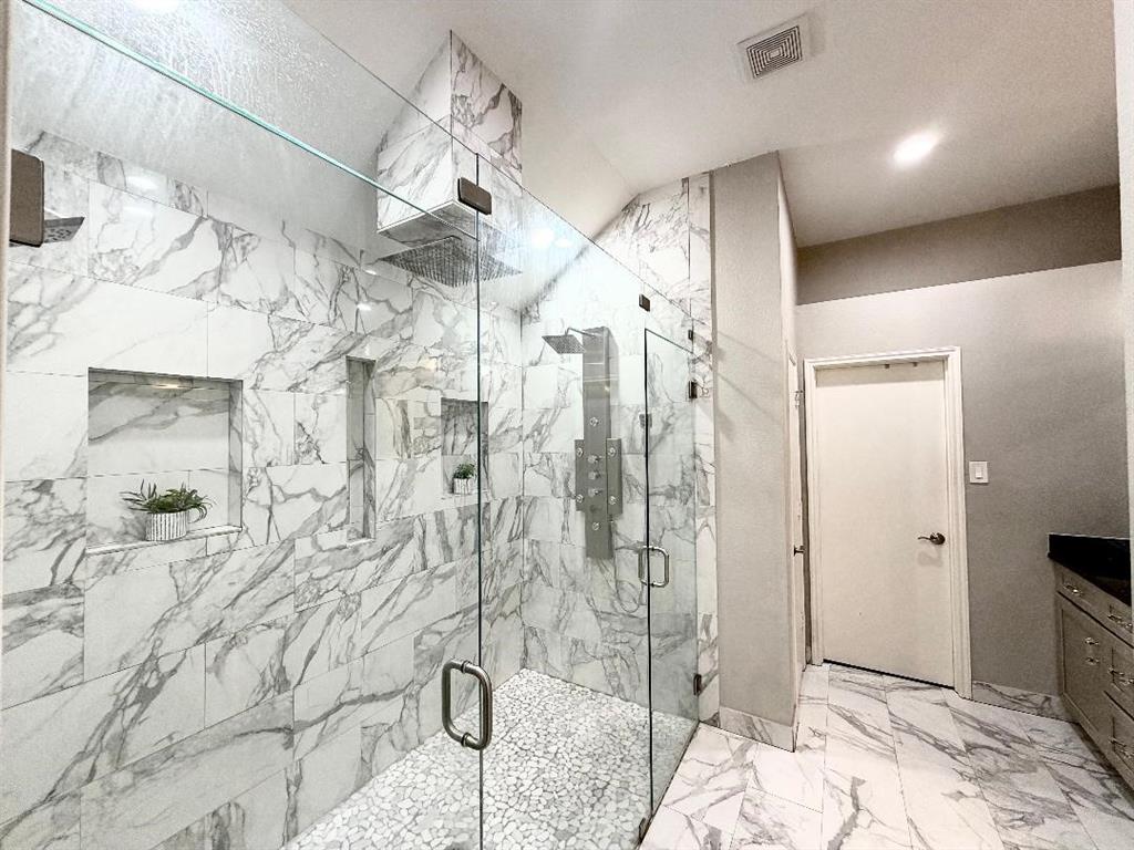 Walk-in shower featuring large-format stone-finish tile surround, pebble-texture flooring, frameless glass enclosure, multiple built-in shower niches, and an integrated multi-head shower system