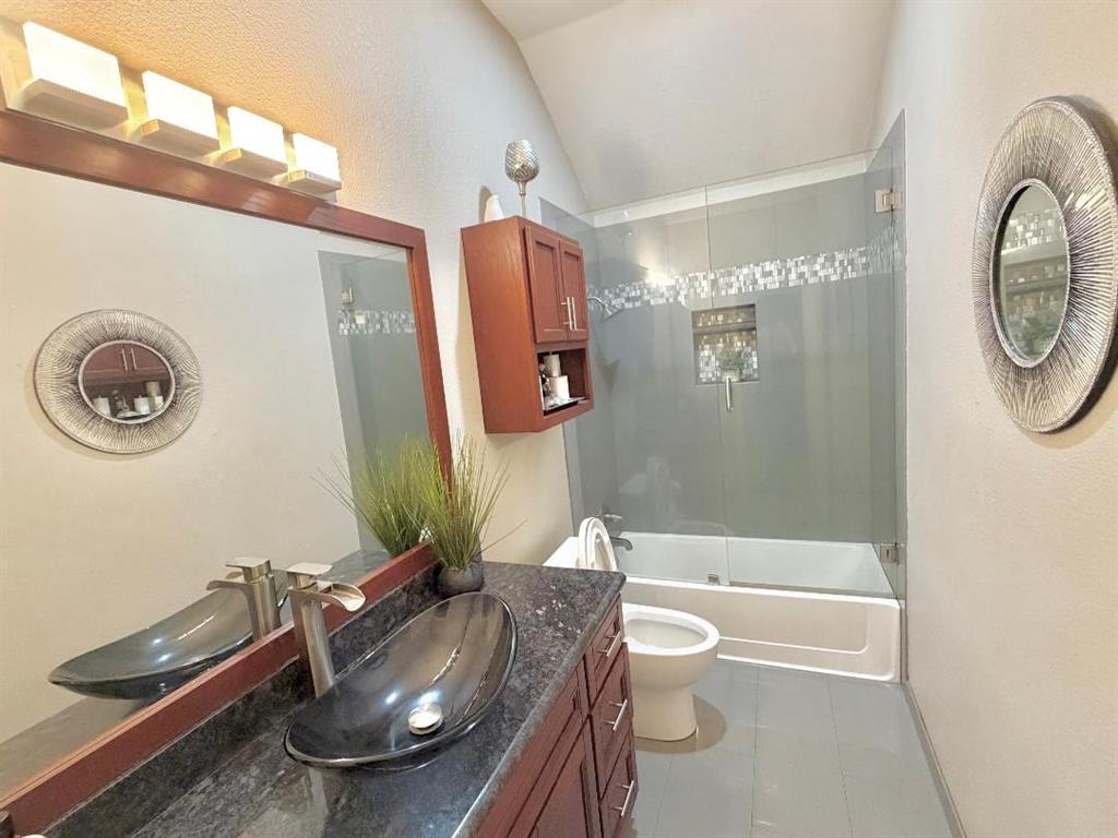 Bathroom featuring a vessel sink with a waterfall faucet, dark stone-finish countertop, wood-finish cabinetry, a glass-enclosed shower, and a vaulted ceiling