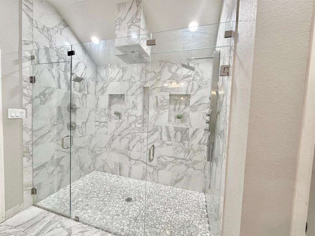 Walk-in shower featuring floor-to-ceiling stone-pattern tile, frameless glass enclosure, pebble-stone flooring, dual recessed wall niches, and a multi-head shower system