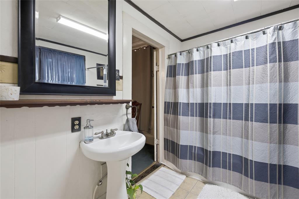 White pedestal sink with a dual-handle faucet, wall-mounted wood-finish shelf, and black-framed mirror