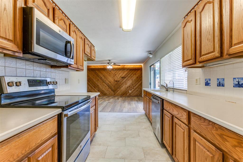 Kitchen featuring oak cabinetry, stainless steel appliances, tile backsplash, and light-toned tile flooring, opening to a space with wood-paneled walls and wood-finish flooring