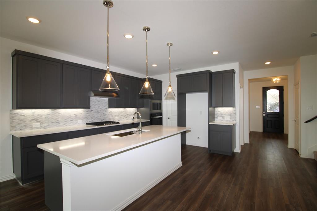Kitchen featuring dark shaker-style cabinetry, quartz countertops, a central island with a sink, glass pendant lighting, and wood-finish flooring
