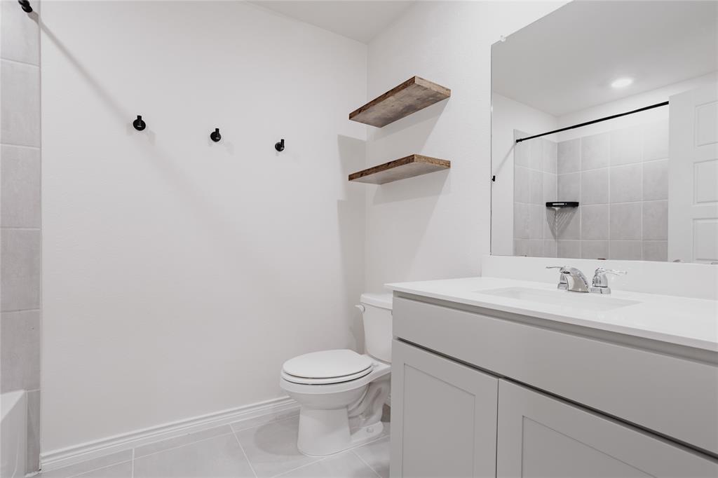 Bathroom featuring a vanity with a white countertop and undermount sink, wall-mounted wood-finish floating shelves, a large frameless mirror, and a tiled shower surround