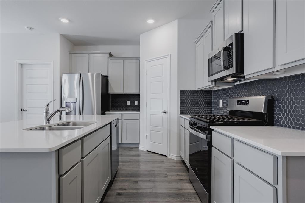 Kitchen featuring light gray shaker-style cabinetry, quartz countertops, a dark hexagonal tile backsplash, stainless steel appliances, and wood-finish flooring