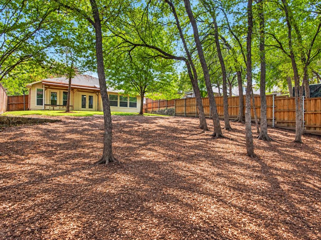 Expansive backyard featuring mature trees, wood privacy fencing, a natural stone retaining wall, and a covered patio