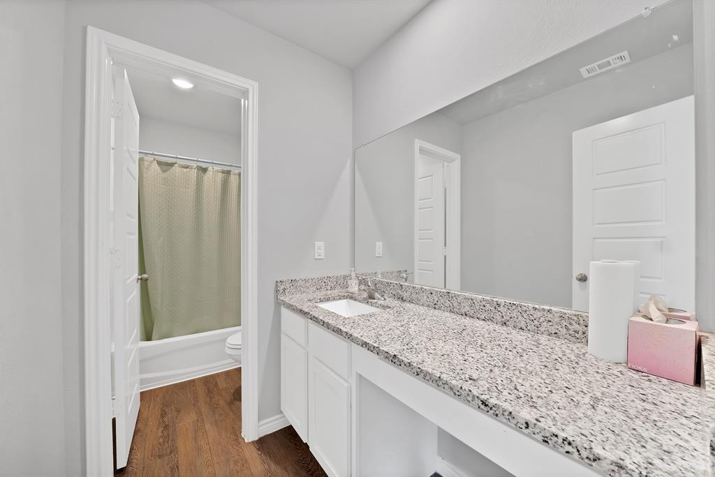 Bathroom vanity featuring white cabinetry, a speckled stone countertop, integrated sink, and full-width wall mirror, with wood-finish flooring