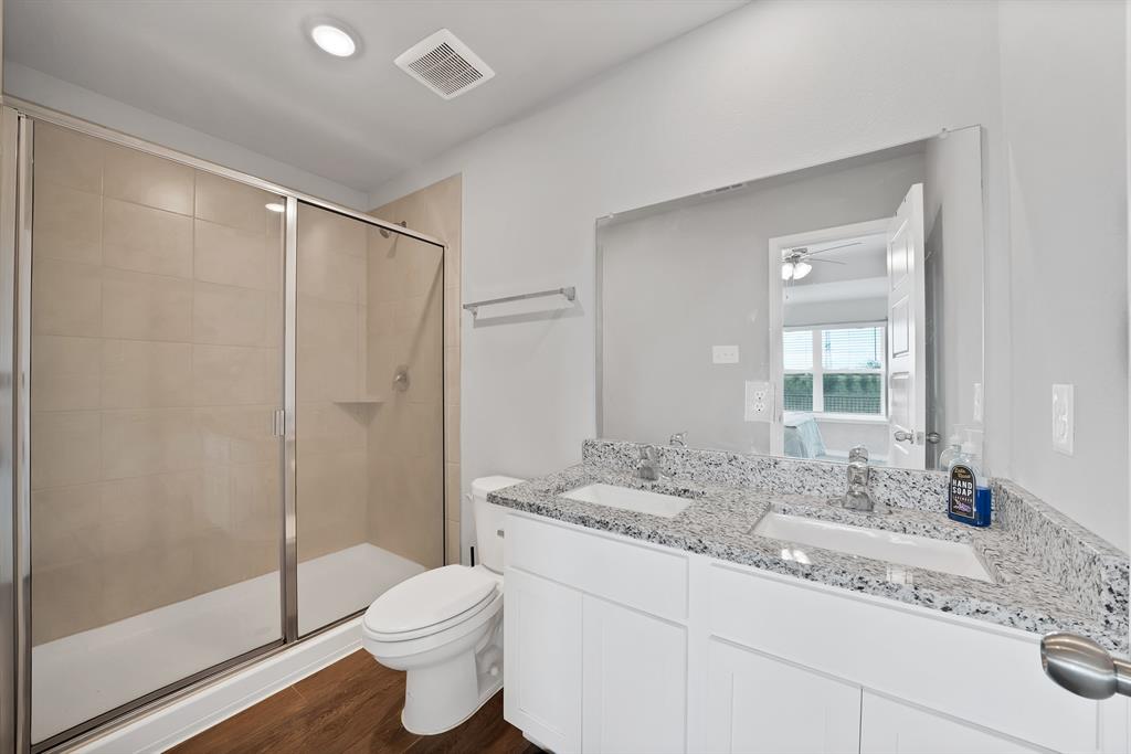 Bathroom featuring a double-sink vanity with light-colored stone-finish countertops, a shower with a sliding glass door and tiled surround, and wood-finish flooring