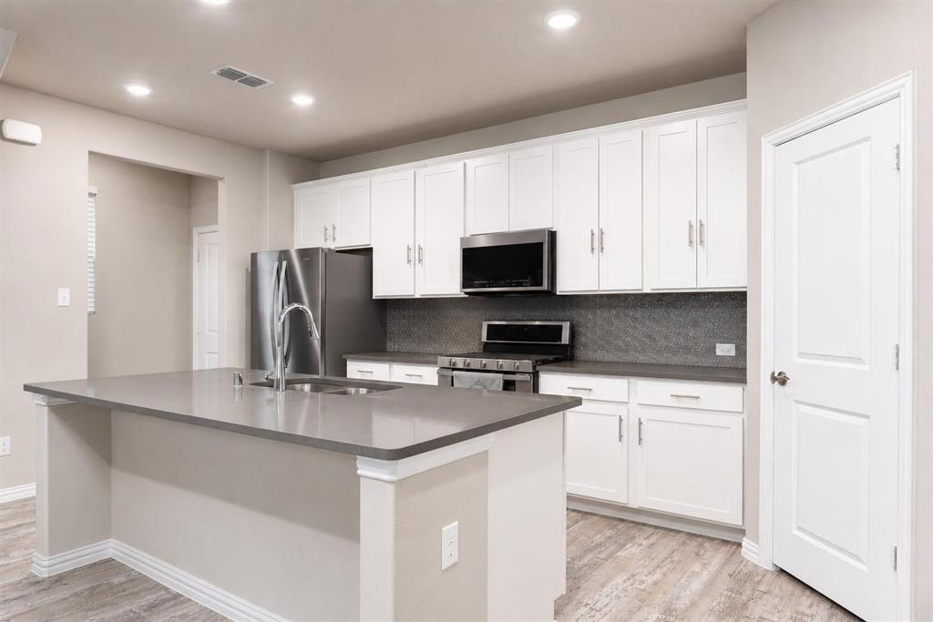 Kitchen featuring white shaker-style cabinetry, quartz countertops, a central island with an integrated sink, stainless steel appliances, and wood-finish flooring