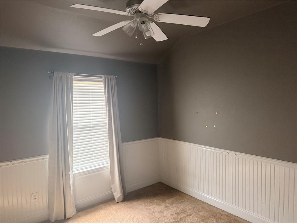 Room featuring neutral wall tones, white beadboard wainscoting, carpeted flooring, a multi-blade ceiling fan, and a single window with blinds Room featuring neutral wall tones, white beadboard wainscoting, carpeted flooring, a multi-blade ceiling fan, and a single window with blinds