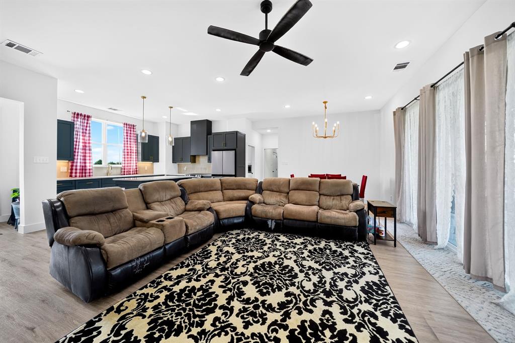 Open-concept living space featuring wood-finish flooring, recessed lighting, matte black ceiling fan, and a kitchen with dark cabinetry and dual pendant lights Open-concept living space featuring wood-finish flooring, recessed lighting, matte black ceiling fan, and a kitchen with dark cabinetry and dual pendant lights