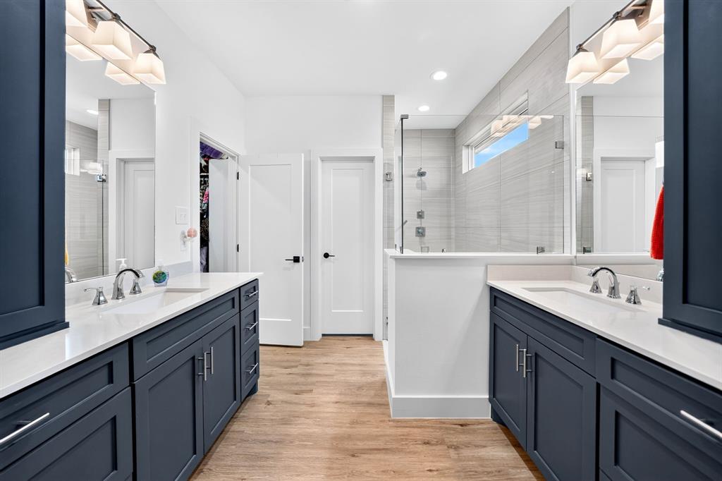 Dual navy cabinetry vanities with light-toned stone countertops, wood-finish flooring, glass-enclosed shower with horizontal tile surround, and polished metal hardware