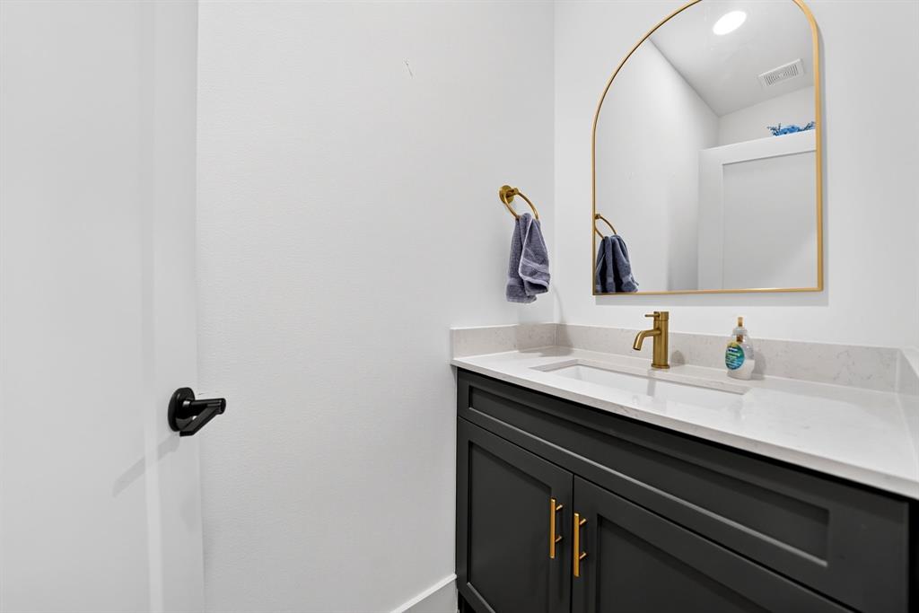 Dark-toned cabinetry featuring gold-finish hardware, quartz countertop, integrated rectangular sink, brass-tone faucet, and arched mirror