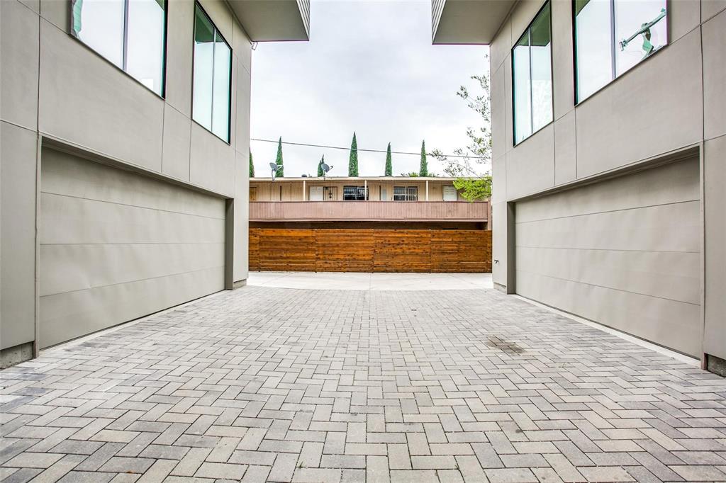 Modern exterior facade featuring smooth-finish cladding, dual flush-mount garage doors, and herringbone-pattern paver motor court enclosed by a horizontal wood privacy fence