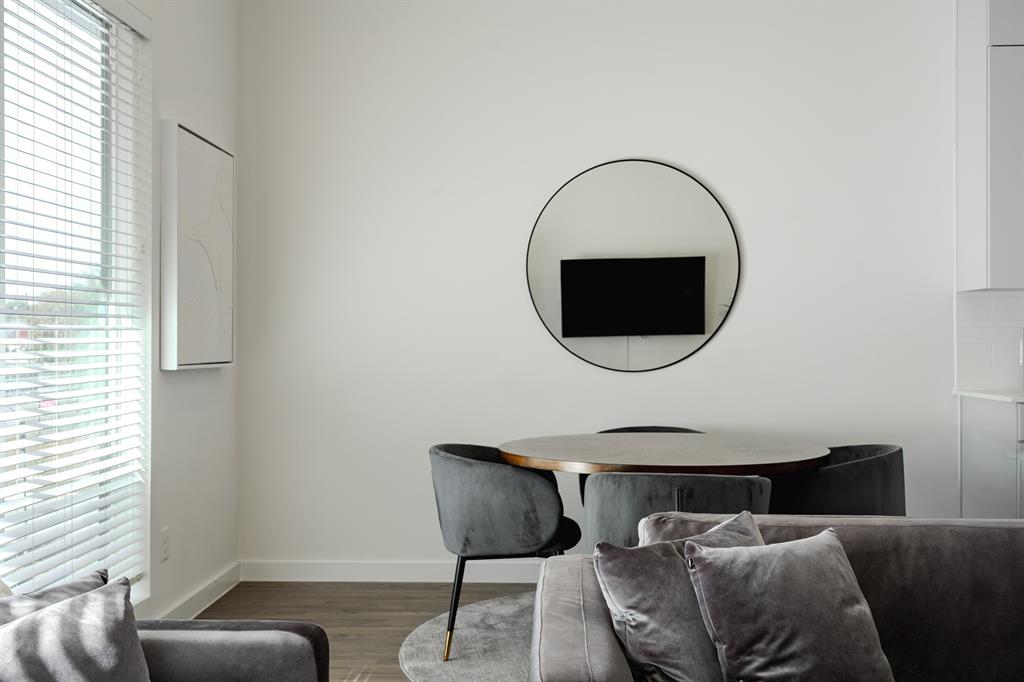 High ceilings, large window with horizontal blinds, wood-finish flooring, and a circular wall-mounted mirror