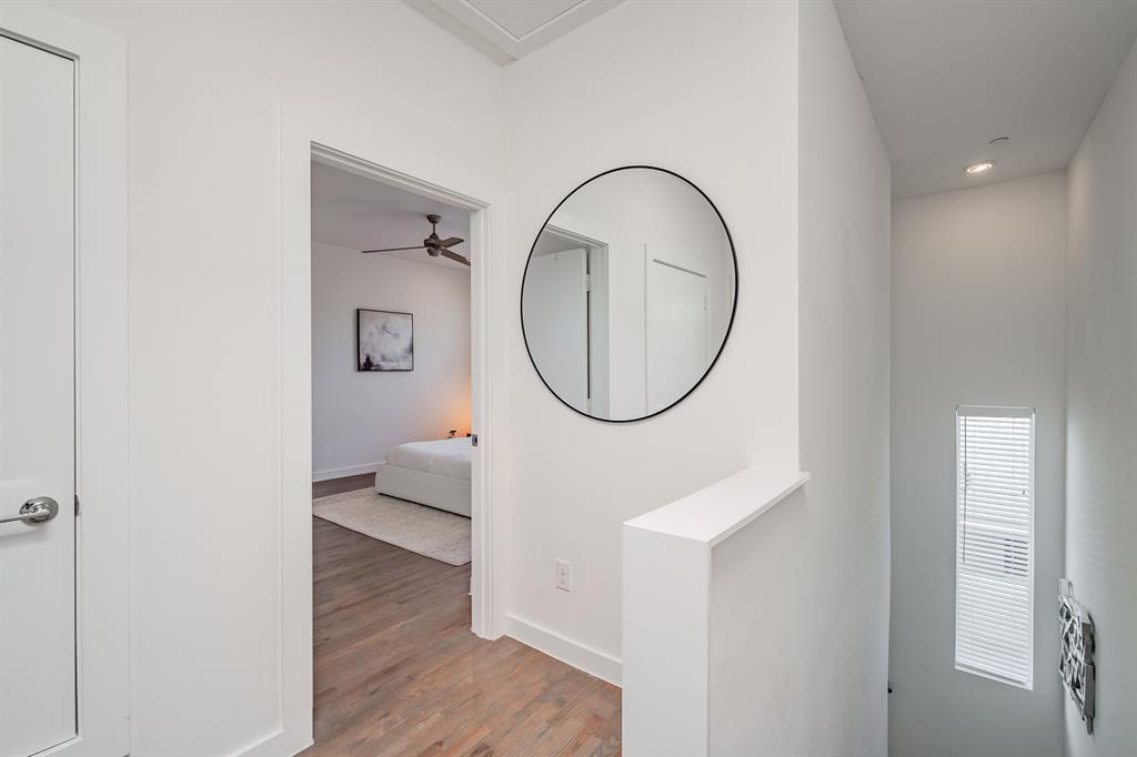 Interior hallway featuring wood-finish flooring, a wall-mounted circular mirror with a black frame, and a tall vertical window with integrated blinds