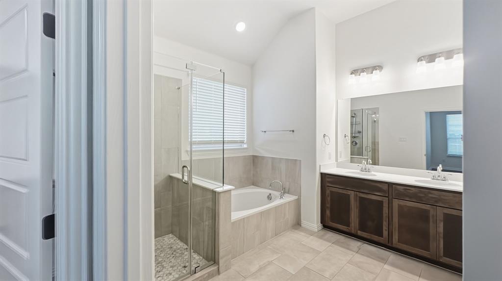 Bathroom featuring a dual-sink vanity with dark-finish cabinetry, a large frameless mirror, a deep soaking tub, a tiled walk-in shower with a glass enclosure, and high vaulted ceilings
