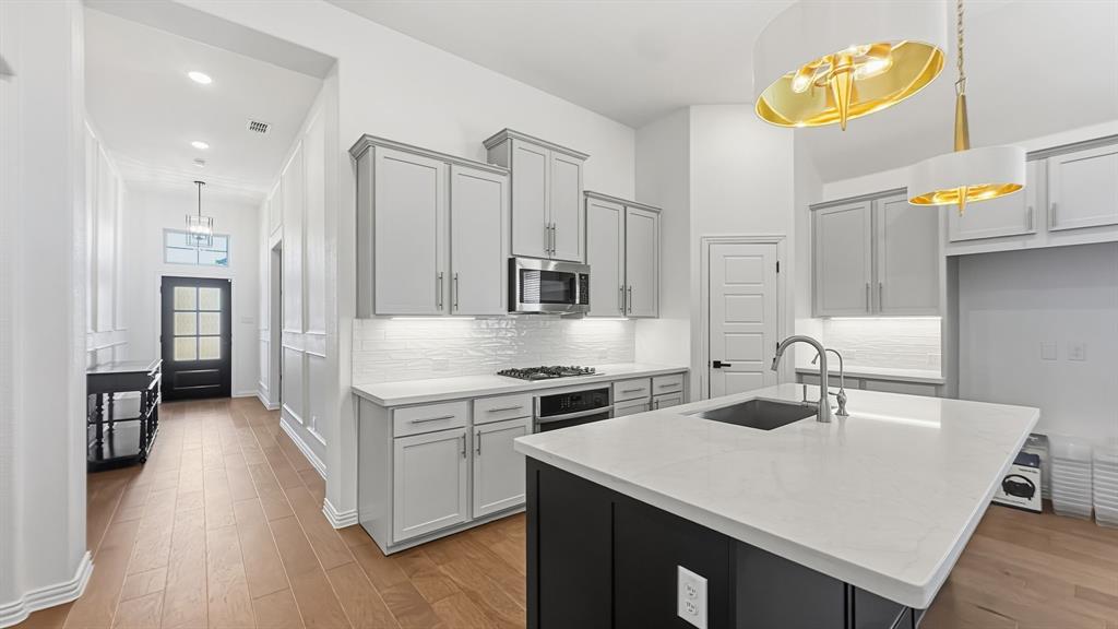 Kitchen featuring light-gray shaker-style cabinetry, white stone countertops, a dark-toned center island with an undermount sink, under-cabinet lighting, and wood-finish flooring