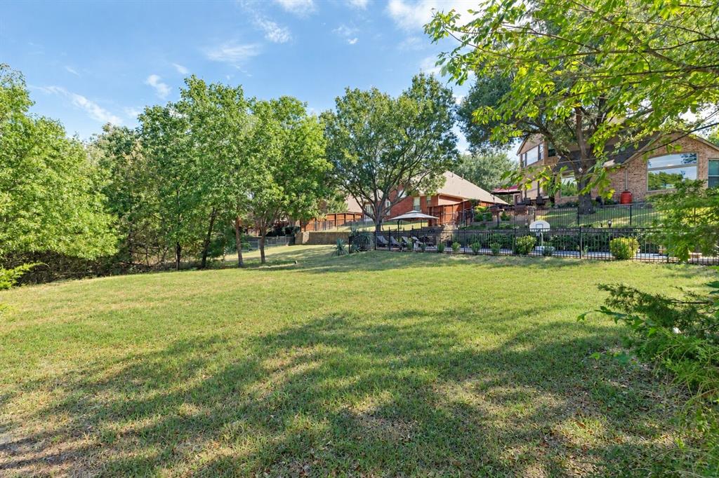 Expansive backyard featuring a manicured lawn, mature trees, tiered stone retaining walls, and a perimeter metal fence