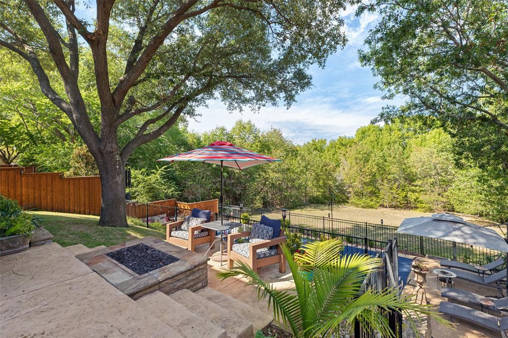 Tiered stone patio featuring a built-in fire pit, metal perimeter fencing, mature trees, and landscaping