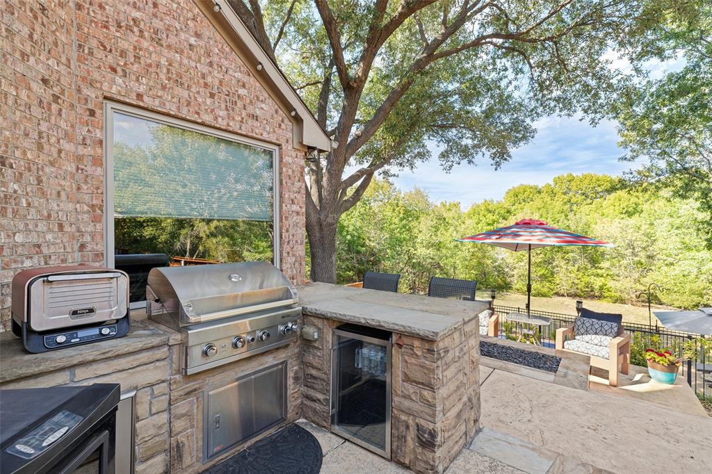 Outdoor kitchen featuring stone masonry, stainless steel built-in grill, under-counter refrigeration, and stone countertops, set against a backdrop of mature trees