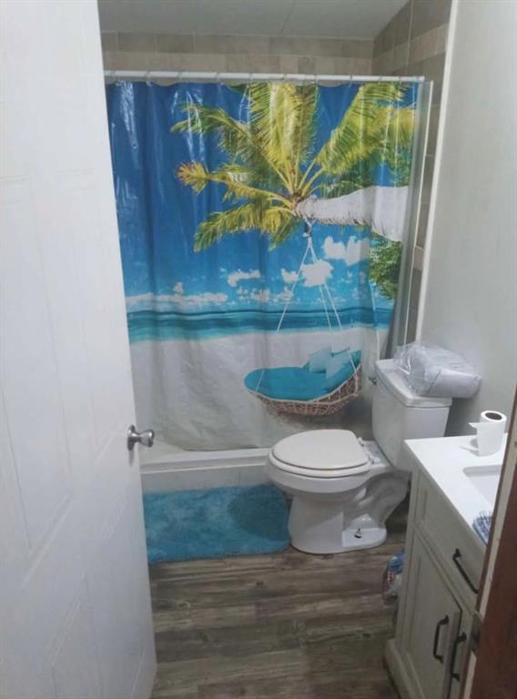 Bathroom featuring wood-finish flooring, a white vanity with dark hardware, a standard toilet, and a tiled shower surround