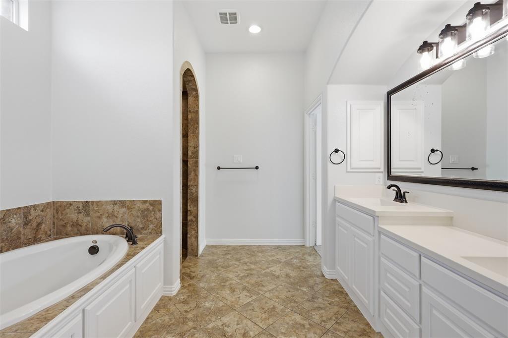 Bathroom featuring a dual-sink vanity with white cabinetry, quartz countertop, framed wall mirror, tiled bathtub surround, and arched tiled shower entry
