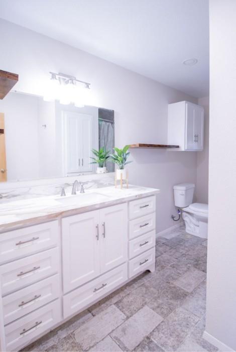 Bathroom featuring a white double-door vanity with stone-finish countertop, undermount sink, multi-drawer storage, and natural stone-finish floor tiling, complemented by white wall-mounted cabinetry and a floating wood-finish shelf