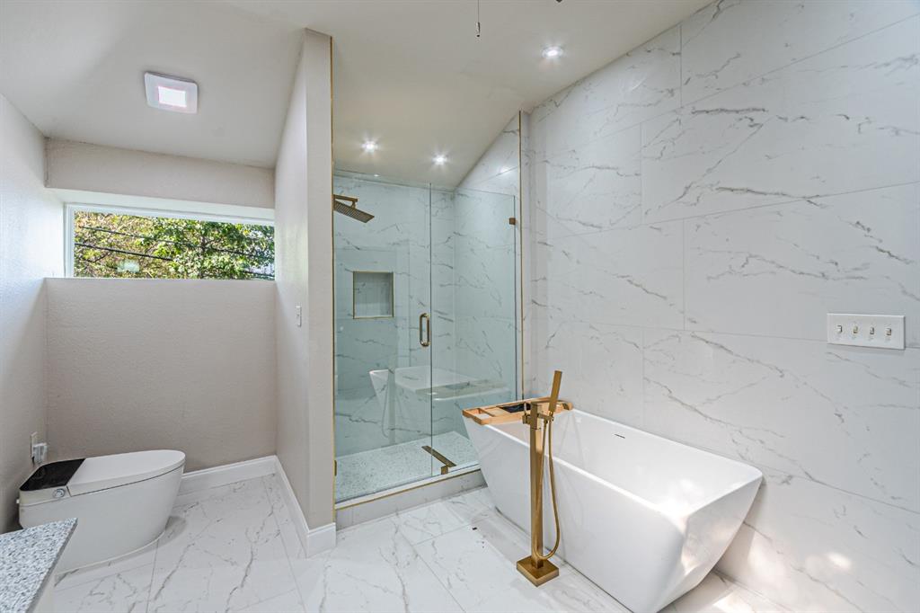 Freestanding soaking tub with a floor-mounted brass faucet, walk-in glass-enclosed shower, built-in shower niche, and large-format tile flooring and wall surfacing