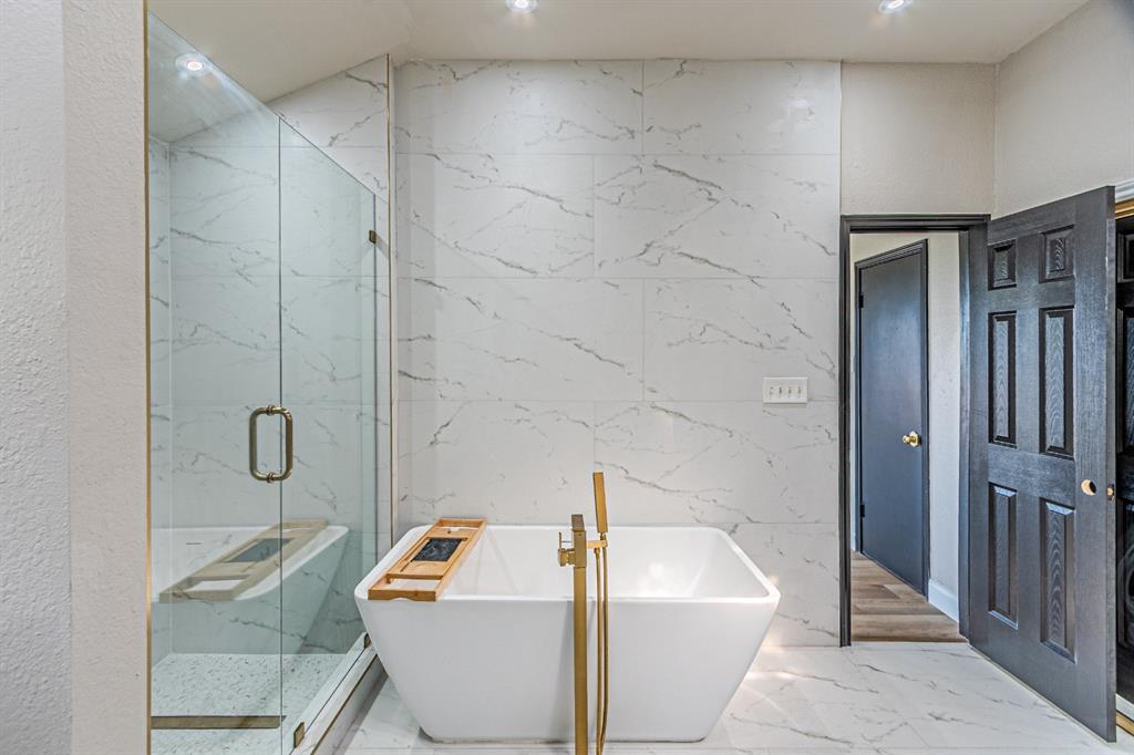 Freestanding soaking tub with brass floor-mounted filler, glass-enclosed walk-in shower, and stone-finish wall tile