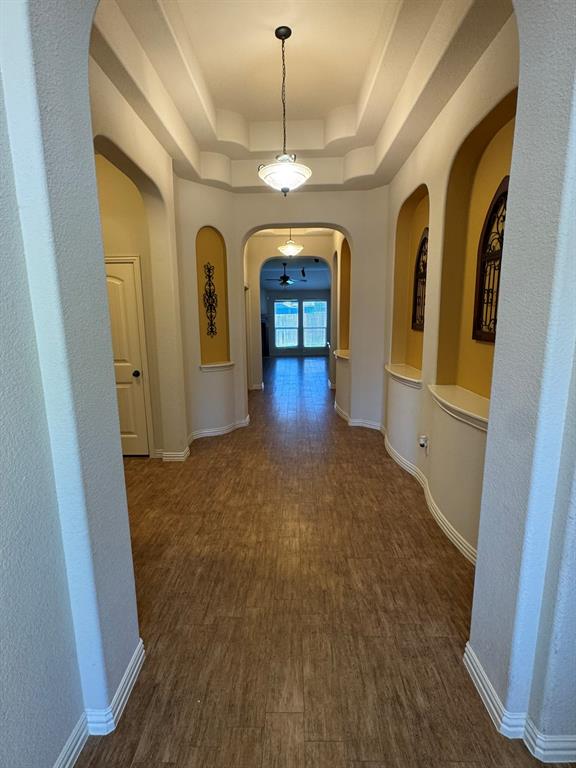 Hallway featuring arched architectural openings, built-in display niches, a tiered tray ceiling, pendant lighting, and wood-finish flooring