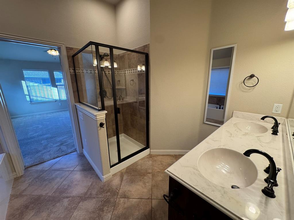 Bathroom featuring dual-sink vanity with stone-finish countertop, dark metal fixtures, tiled flooring, and a walk-in shower with a black-frame enclosure